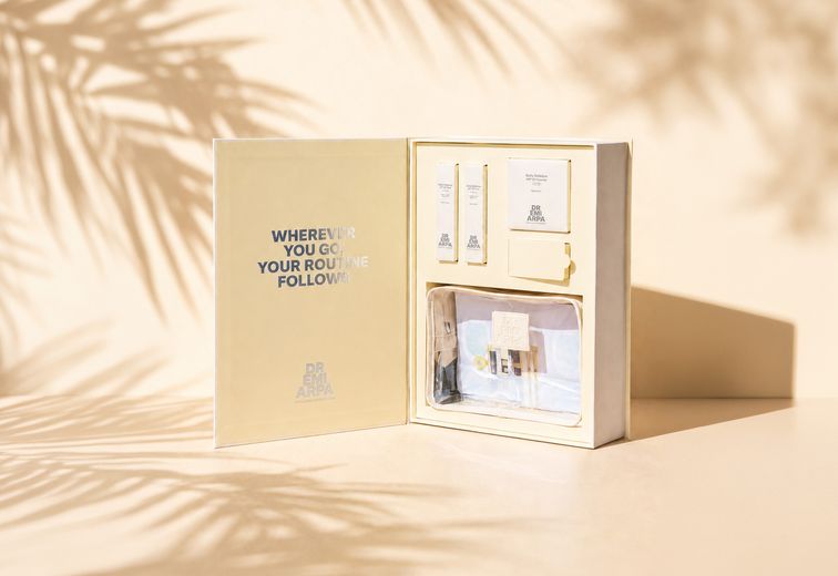 Offene Hautpflege-Set-Box mit verschiedenen Produkten darin, beschriftet mit „WHEREVER YOU GO, YOUR ROUTINE FOLLOWS“. Deutlicher Schatten von Palmblättern.