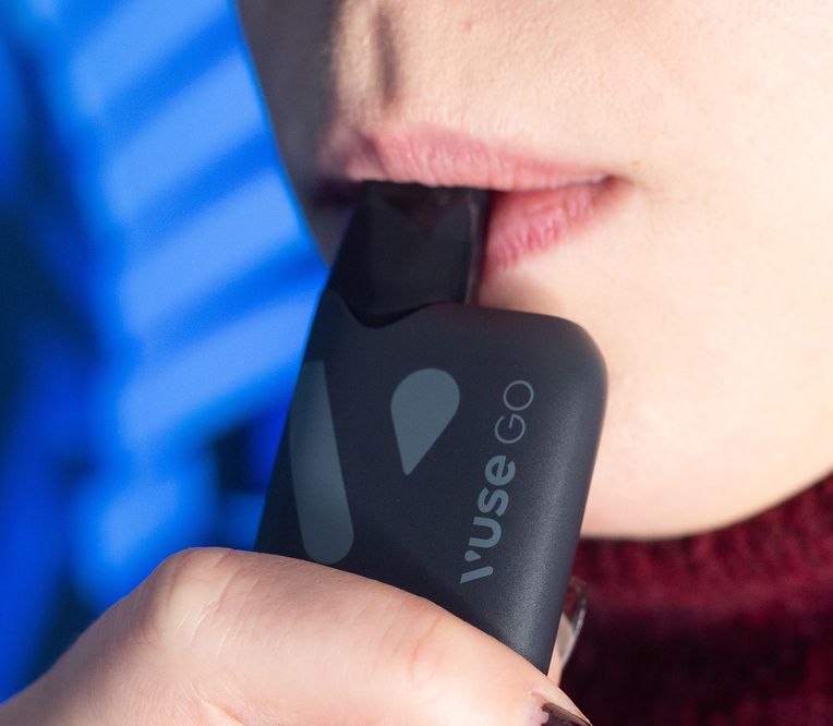 Nahaufnahme einer Person, die einen Vuse Go Vape benutzt, wobei ihre Lippen das schwarze Gerät berühren. Der Hintergrund hat einen blauen Farbton.