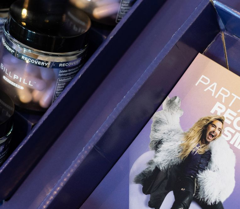 Dosen mit Erholungspillen in einer Ausstellungsbox mit einer Broschüre, auf der eine Person in einem flauschigen Mantel zu sehen ist, die lebhaft gestikuliert, begleitet von dem Text „Party Hard, Recover Smart“.