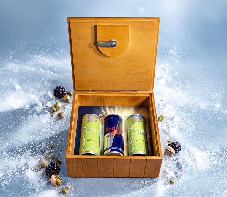 Offene Holzkiste mit zwei Energy-Drinks mit Pistaziengeschmack und einer Red-Bull-Dose, umgeben von Eis, Pistazien und Brombeeren auf einer vereisten Oberfläche.