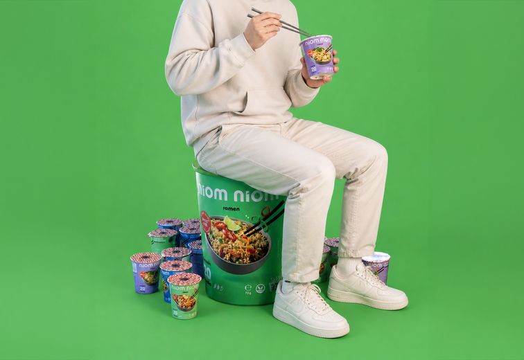 Person in beige outfit sits on a giant ramen cup holding smaller noodle cup with chopsticks, surrounded by more cups on green background.