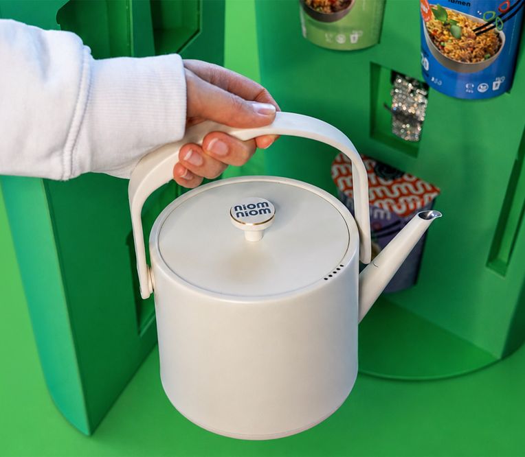 Eine Hand hält eine weiße Teekanne mit der Aufschrift „niom niom“ vor einem grünen Automaten, in dem Lebensmittel ausgestellt sind.