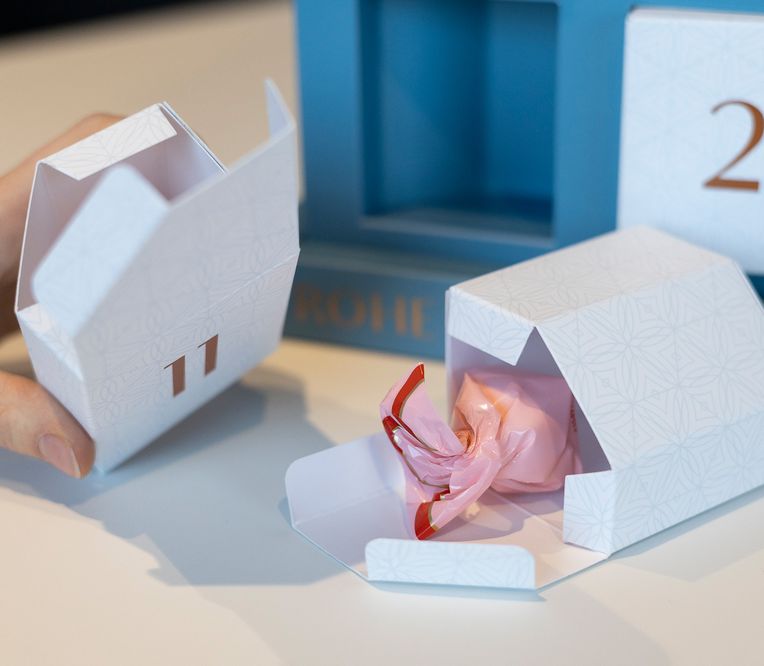 Eine Hand öffnet eine Adventskalender-Schachtel mit den Zahlen 11 und 2 und enthüllt darin einen in rosa Papier eingewickelten Gegenstand.