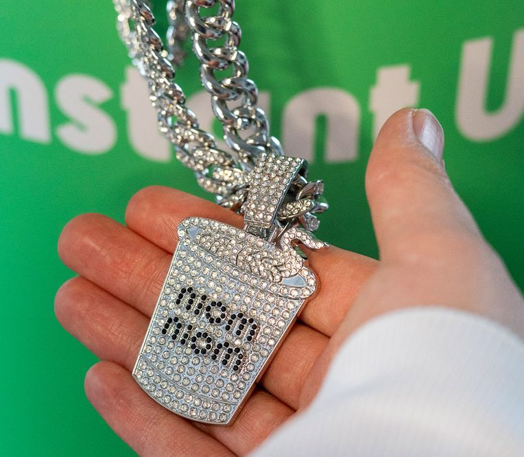 Eine Hand hält eine funkelnde Kette mit einem Anhänger in Form eines Nudelbechers, auf dem vor grünem Hintergrund „niom niom“ steht.