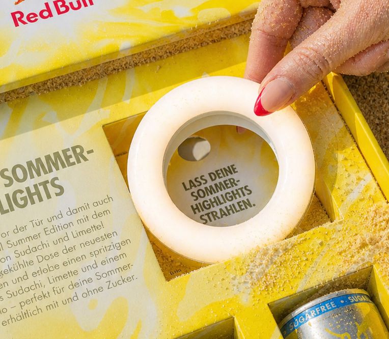 Nahaufnahme einer sandigen Hand, die eine runde Leuchte in einer farbenfrohen Red-Bull-Verpackung hält, mit deutschem Text über die Sommer-Highlights.