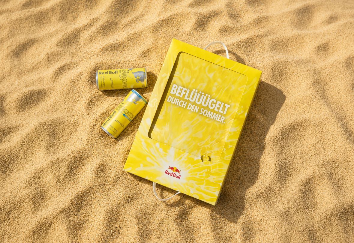 Gelbe Red-Bull-Dosen und eine dazu passende Tasche mit Schriftzug im Sand, die für eine Sommeredition werben.