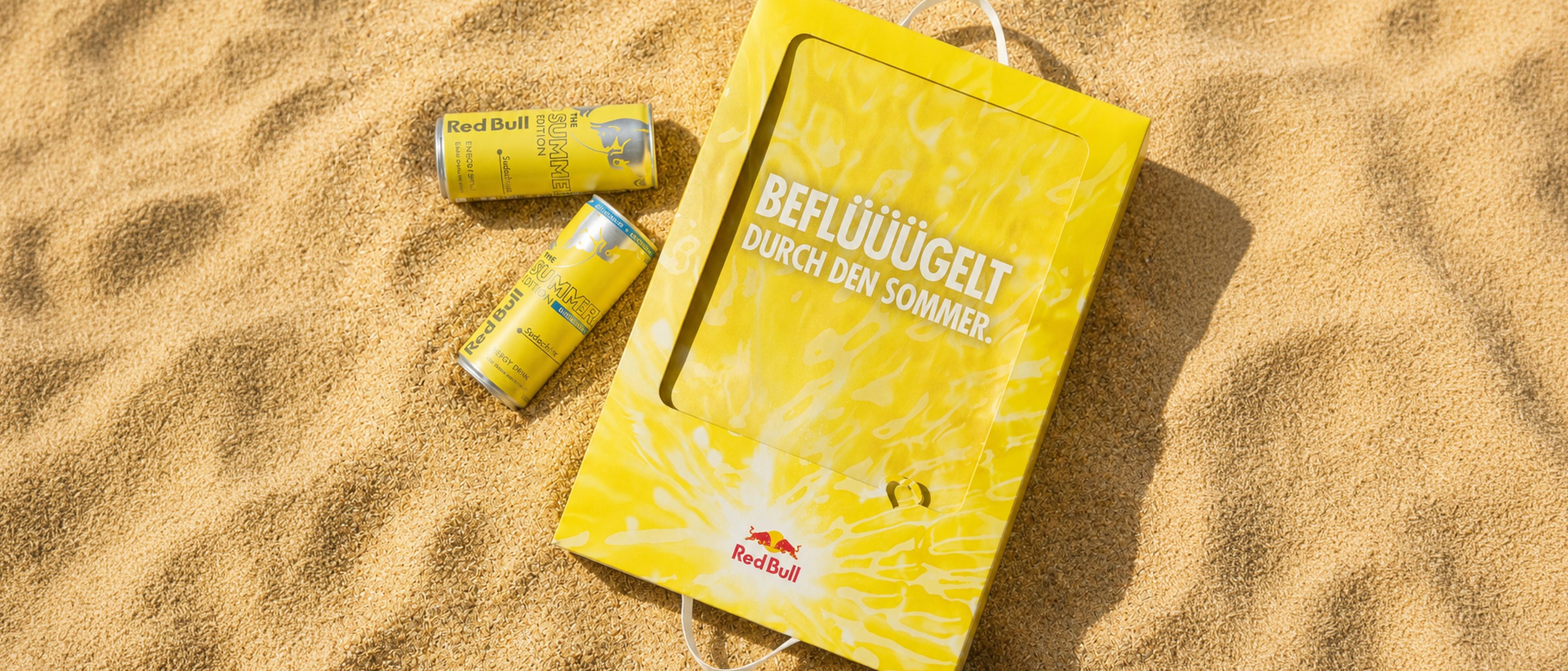 Gelbe Red-Bull-Dosen und eine dazu passende Tasche mit Schriftzug im Sand, die für eine Sommeredition werben.