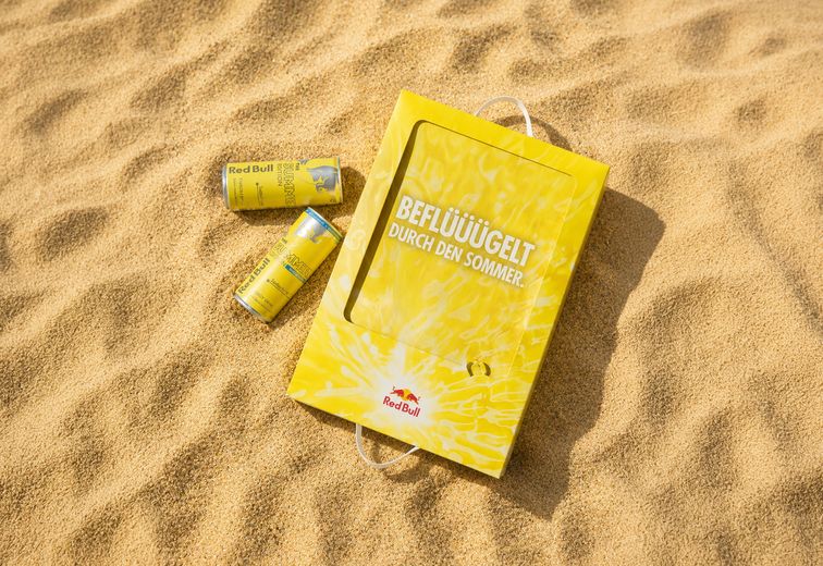 Gelbe Red-Bull-Dosen und eine dazu passende Tasche mit Schriftzug im Sand, die für eine Sommeredition werben.