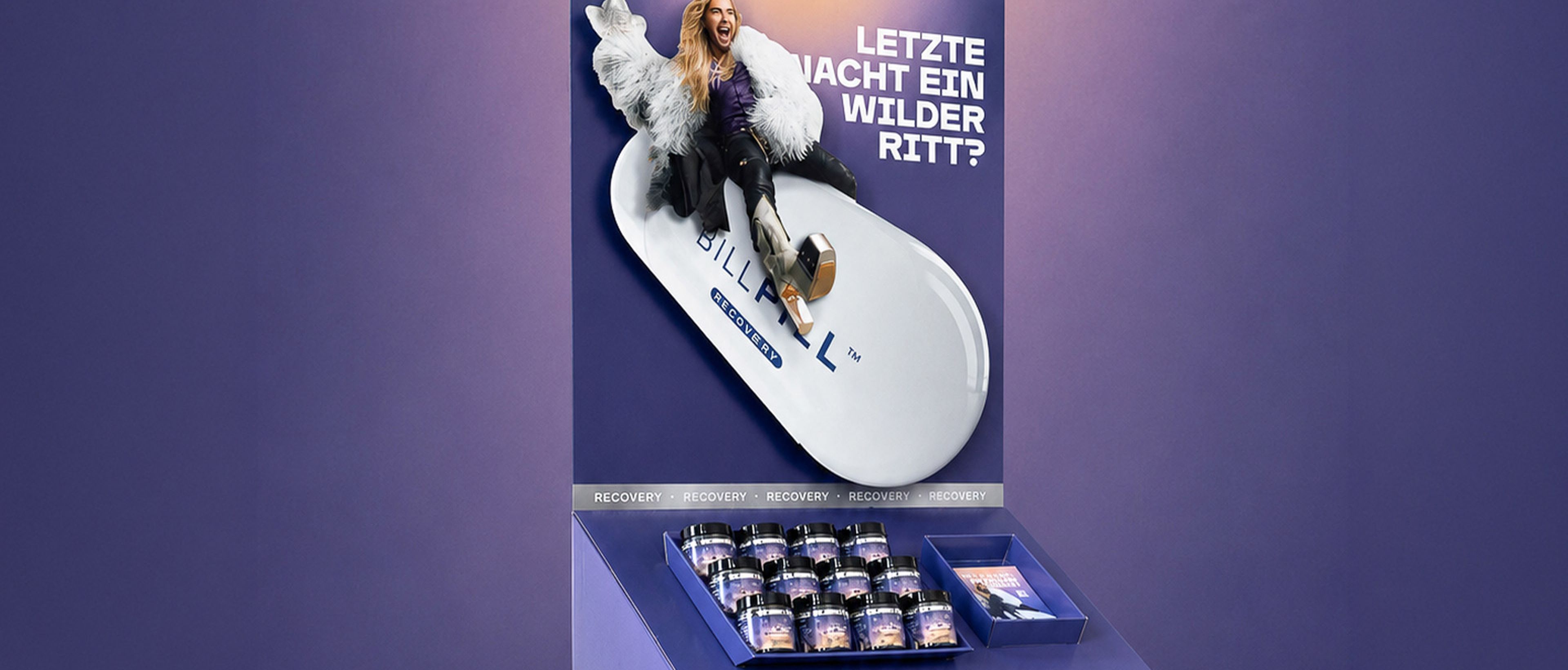 Ein violetter Ausstellungsständer in Form einer riesigen Pille, mit Flaschen und einem Kartenschlitz, der mit einem deutschen Text für „BILLPILL Recovery“ wirbt.
