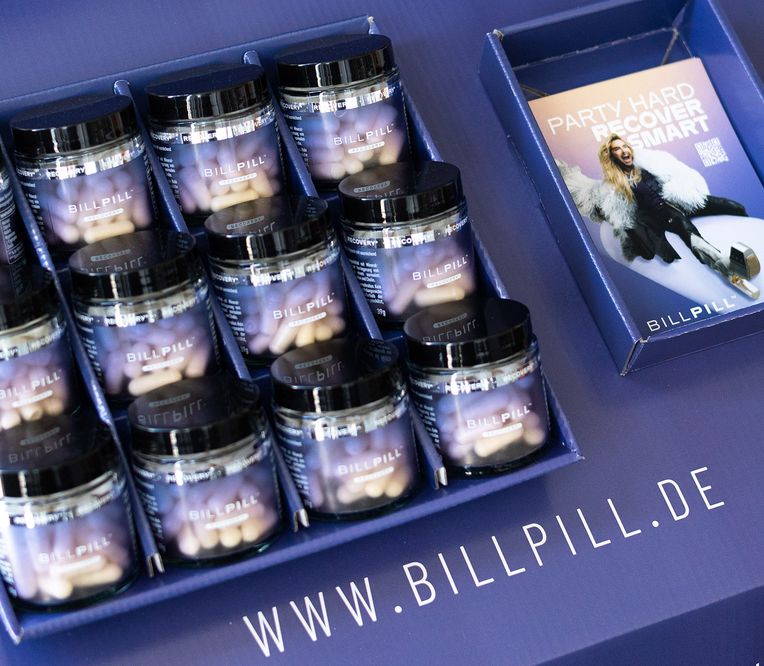 Auslage mehrerer Gläser mit der Aufschrift „BILLPILL“, die Kapseln enthalten, zusammen mit Werbematerial mit dem Schriftzug „Party Hard, Recover Smart“.