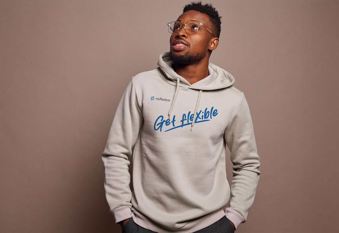 Ein Mann mit Brille und einem beigen Kapuzenpulli mit der Aufschrift „Get flexible“, der vor einem einfarbigen Hintergrund nach oben blickt.