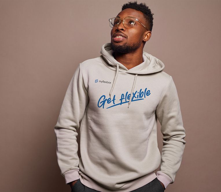 Ein Mann mit Brille und einem beigen Kapuzenpulli mit der Aufschrift „Get flexible“, der vor einem einfarbigen Hintergrund nach oben blickt.