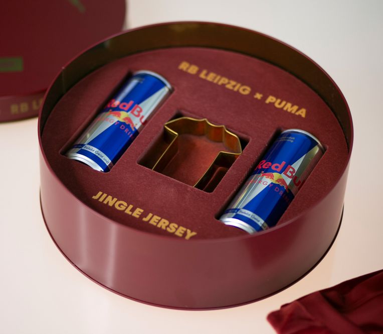 Runde Geschenkbox mit zwei Red-Bull-Dosen und einer Keksausstecher in Trikotform, beschriftet mit „RB Leipzig x Puma“ und „Jingle Jersey“.