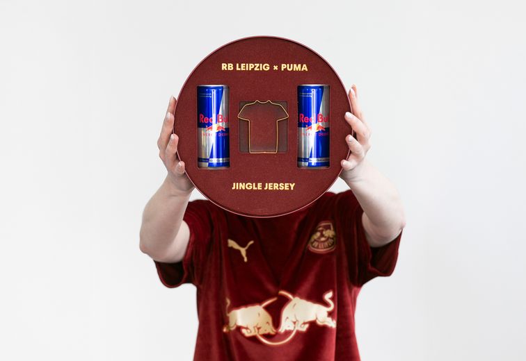 Eine Person, die eine runde RB Leipzig x Puma Jingle Jersey-Box mit zwei Red Bull-Dosen und dem Umriss eines Trikots in der Hand hält.