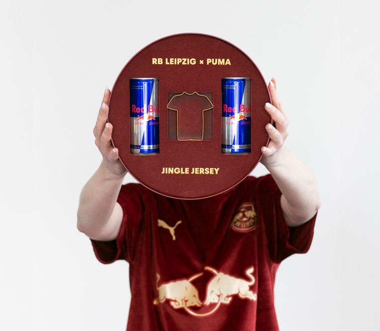 Eine Person, die eine runde RB Leipzig x Puma Jingle Jersey-Box mit zwei Red Bull-Dosen und dem Umriss eines Trikots in der Hand hält.
