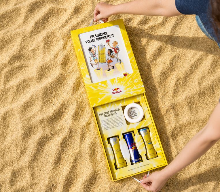 Eine Person öffnet auf dem Sand ein gelbes Red-Bull-Sommer-Set, das zwei Dosen und ein Glas enthält.