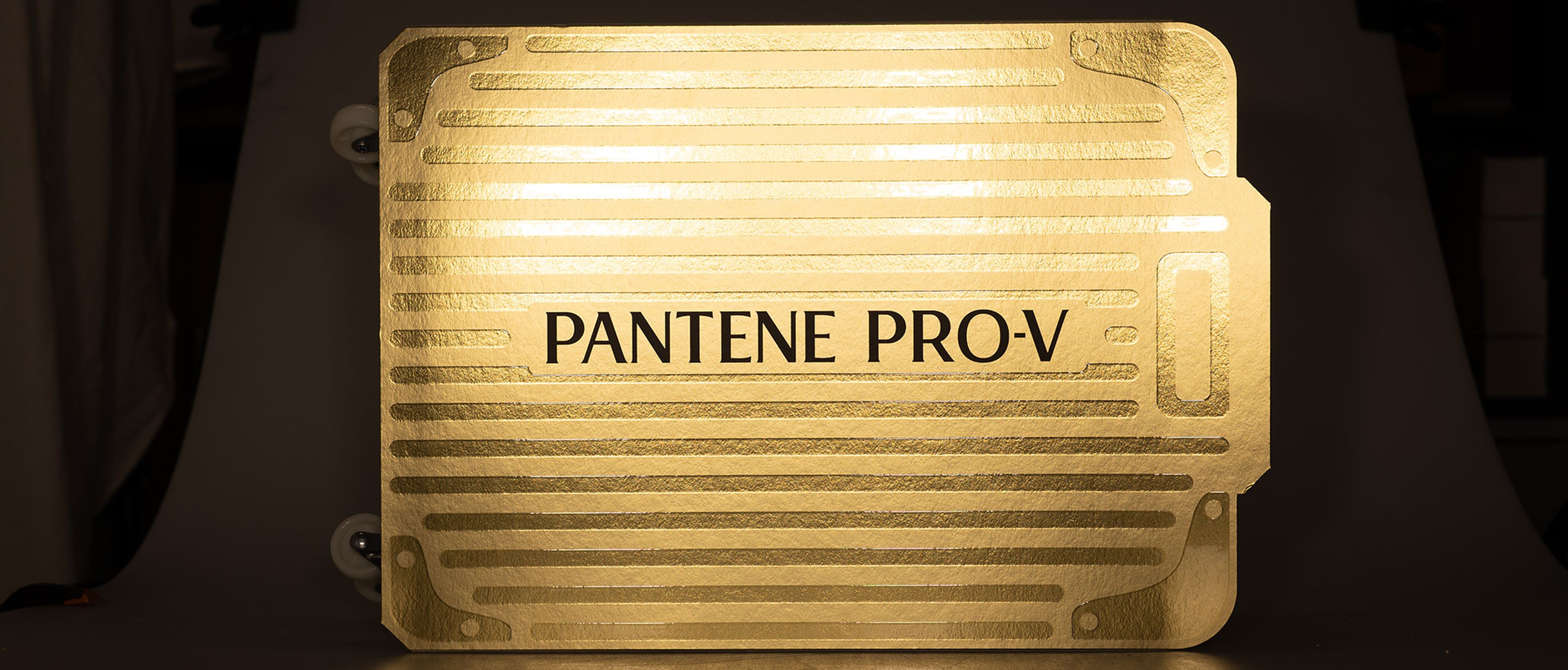Ein leuchtender, metallisch-goldener Koffer mit der Aufschrift „Pantene Pro-V“, das vor einem dunklen Hintergrund beleuchtet ist.