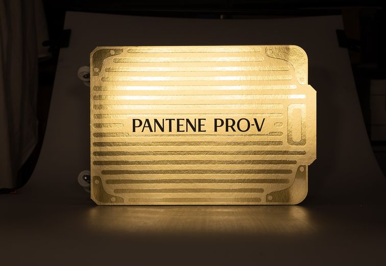 Ein leuchtender, metallisch-goldener Koffer mit der Aufschrift „Pantene Pro-V“, das vor einem dunklen Hintergrund beleuchtet ist.