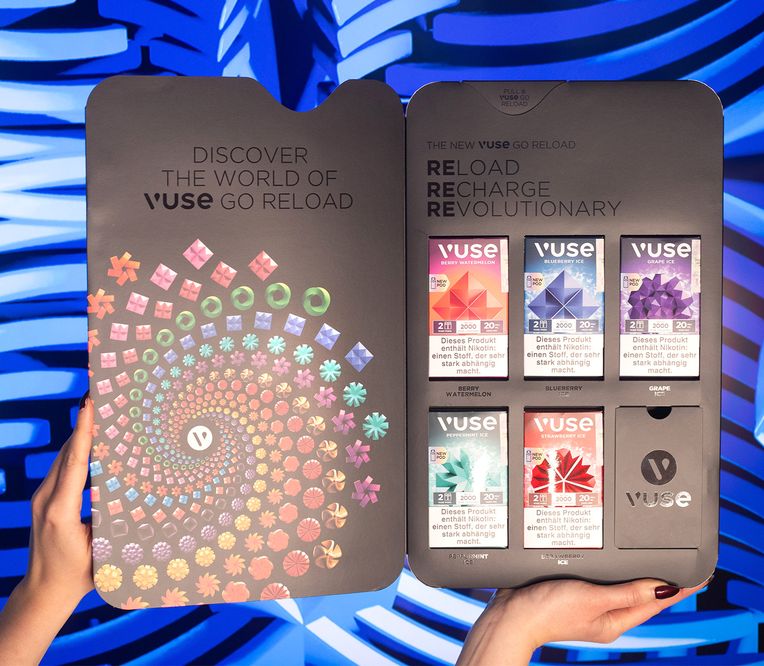 Hände, die eine geöffnete Vuse Go Reload-Verpackung halten, mit farbenfrohem Design und Werbetext auf blau gemustertem Hintergrund.