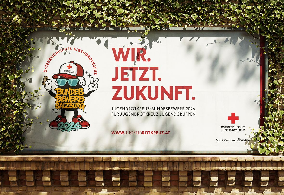 Eine Plakatwand mit der Aufschrift „WIR. JETZT. ZUKUNFT.“, die für den Jugendwettbewerb des Roten Kreuzes in Salzburg 2026 wirbt und von grünem Efeu umrankt ist.