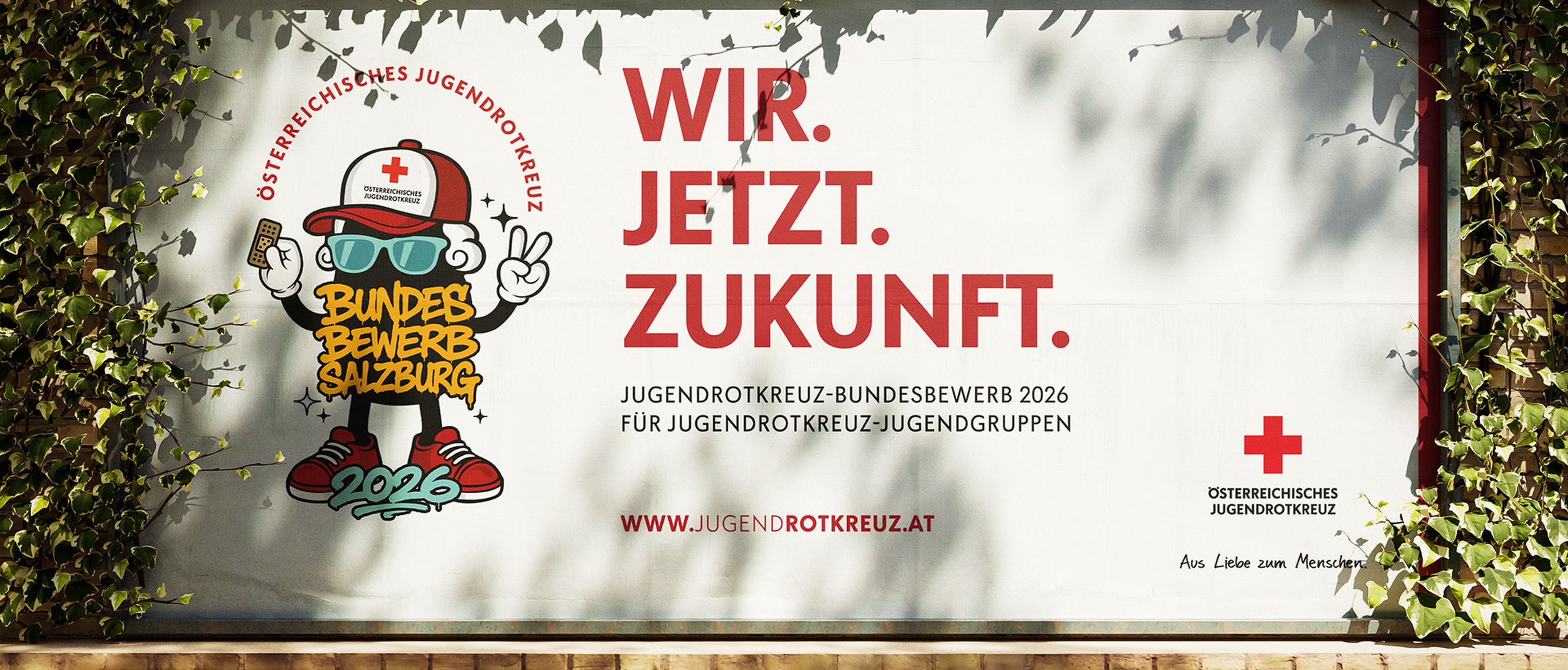 Eine Plakatwand mit der Aufschrift „WIR. JETZT. ZUKUNFT.“, die für den Jugendwettbewerb des Roten Kreuzes in Salzburg 2026 wirbt und von grünem Efeu umrankt ist.
