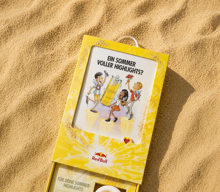 Red-Bull-Verpackung auf Sand mit Comicfiguren, die den Sommer genießen. Der deutsche Text lautet: „Ein Sommer voller Höhepunkte?“