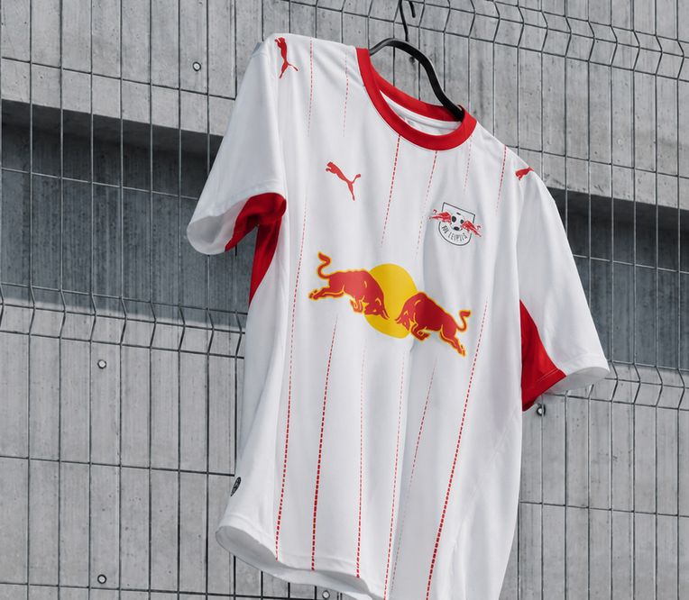 Ein weißes Fußballtrikot mit roten Akzenten und einem Logo, auf dem zwei Stiere abgebildet sind, hängt an einem Drahtzaun vor einer Betonmauer.