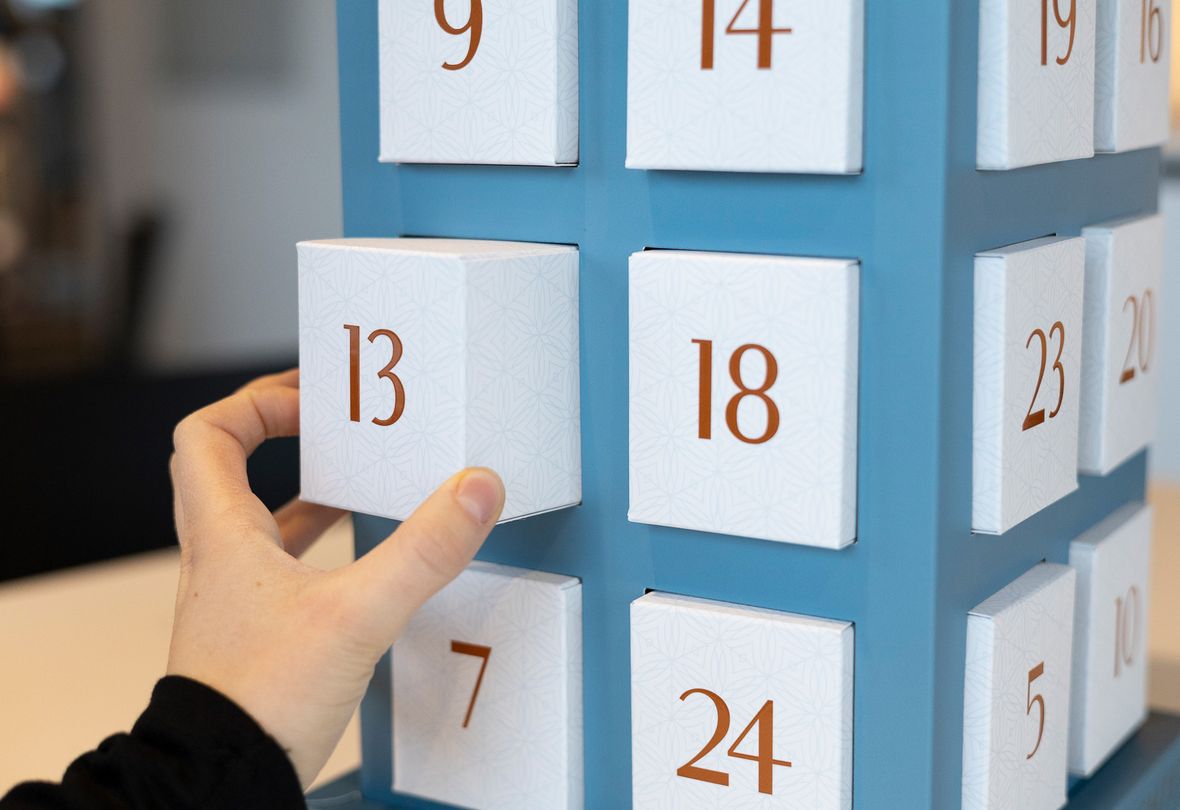 Eine Hand öffnet das Türchen Nr. 13 eines blauen Adventskalenders, daneben sind weitere nummerierte Türchen zu sehen.