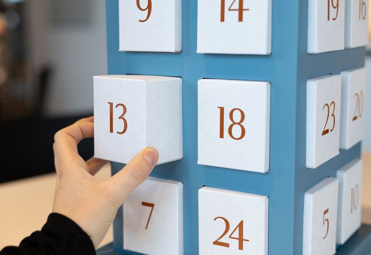 Eine Hand öffnet das Türchen Nr. 13 eines blauen Adventskalenders, daneben sind weitere nummerierte Türchen zu sehen.