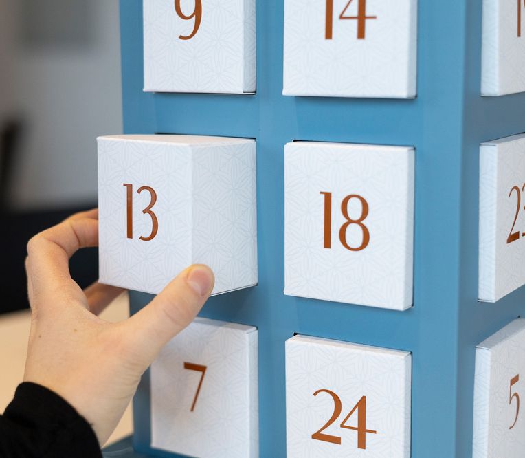 Eine Hand öffnet das Türchen Nr. 13 eines blauen Adventskalenders, daneben sind weitere nummerierte Türchen zu sehen.