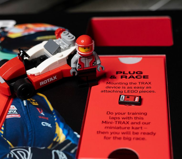 A LEGO figure with a red helmet stands beside a small Rotax kart model on a red "Plug & Race" instructional display.