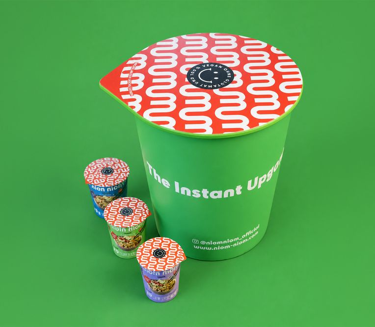 A large green instant noodle cup with smaller colorful cups on a green background, featuring bold patterns and text.