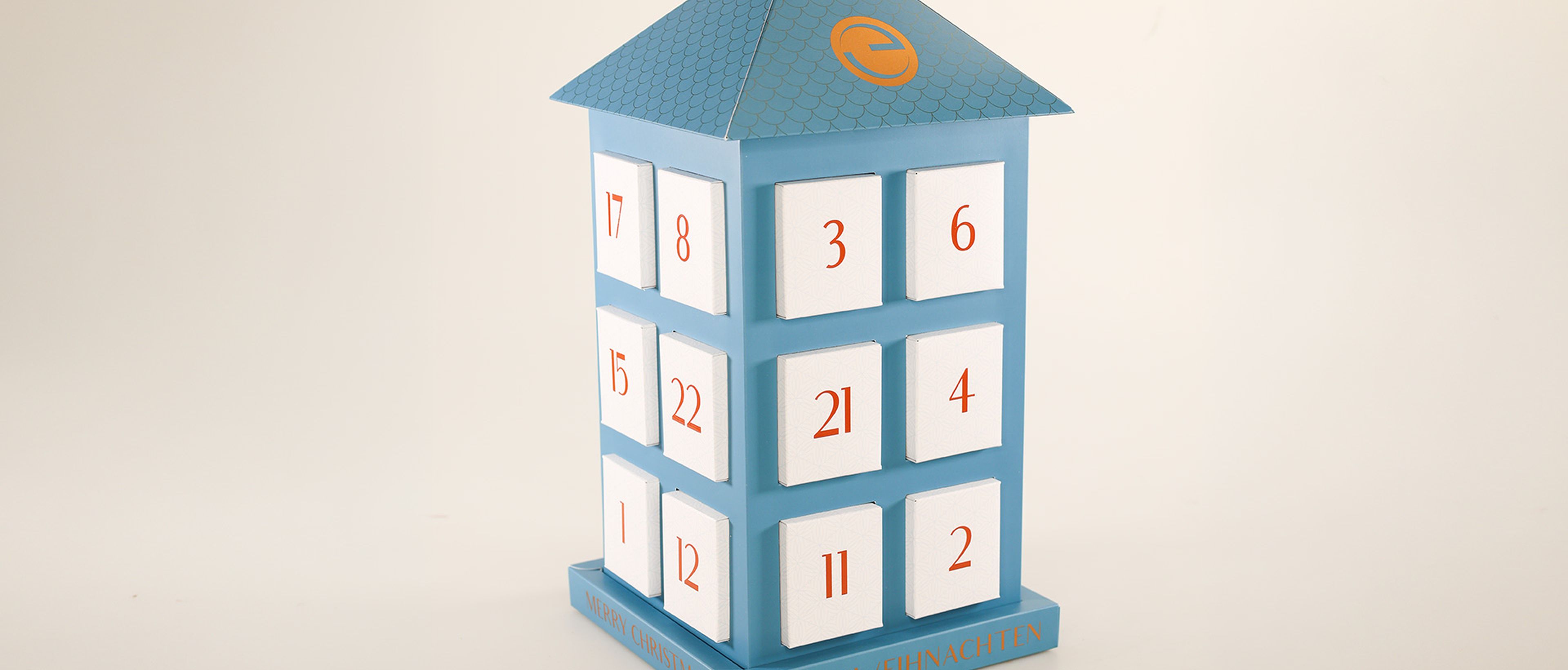 Blauer Adventskalender in Form eines Hauses mit weißen, nummerierten Türchen und der Aufschrift „Frohe Weihnachten“ auf dem Sockel.