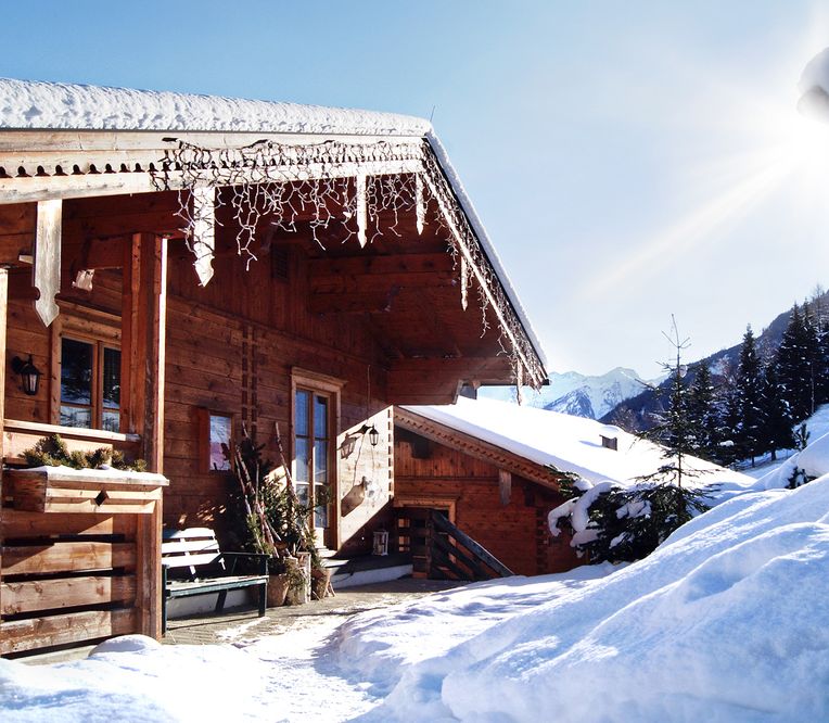 Eine Holzhütte mit schneebedecktem Dach und Eiszapfen, umgeben von Schnee und Kiefern, unter strahlend blauem Himmel und bei strahlendem Sonnenschein.