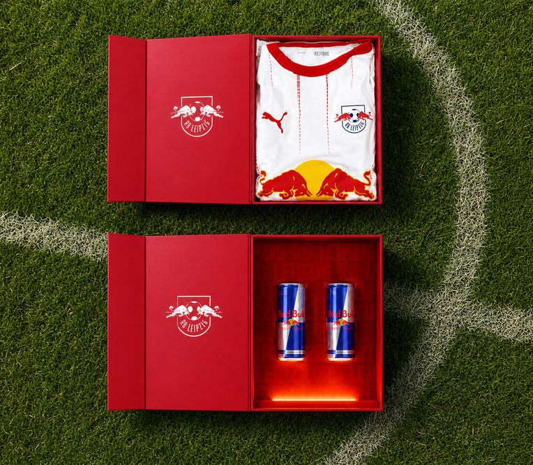 Ein rot-weißes Trikot des RB Leipzig und zwei Red-Bull-Dosen in roten Kartons auf einer Rasenfläche.
