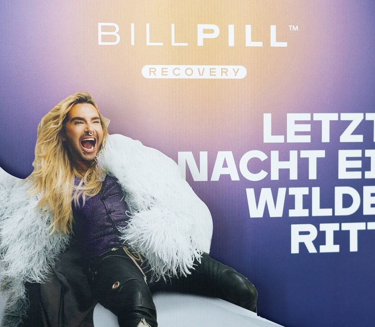 Eine Person mit langen blonden Haaren und einem Nasenring posiert begeistert vor einem violetten Hintergrund, auf dem deutscher Text und die Worte „BillPill Recovery“ zu sehen sind.