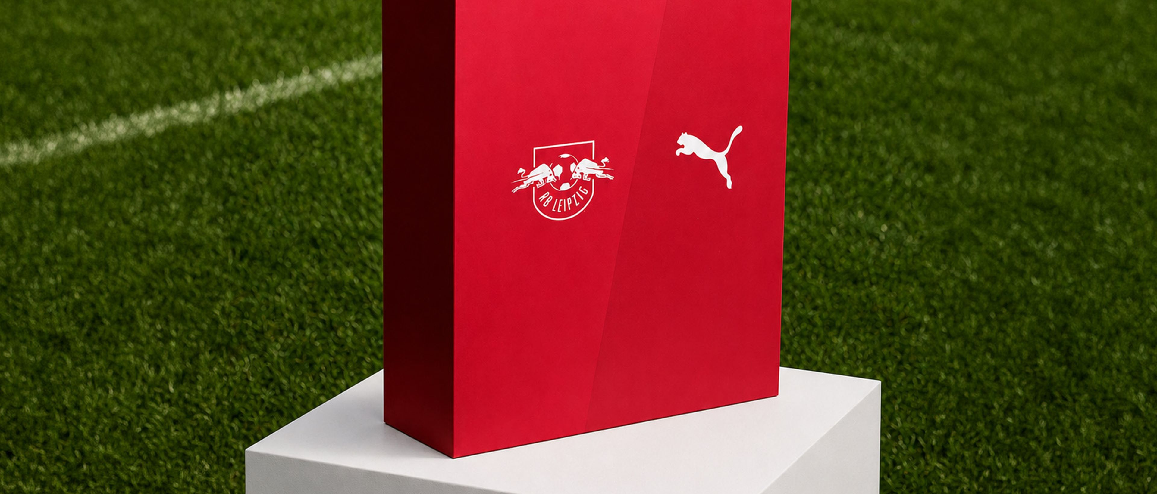 Eine rote Schachtel mit den Logos von RB Leipzig und Puma, die auf einem weißen Sockel steht, im Hintergrund ein Rasenfeld.